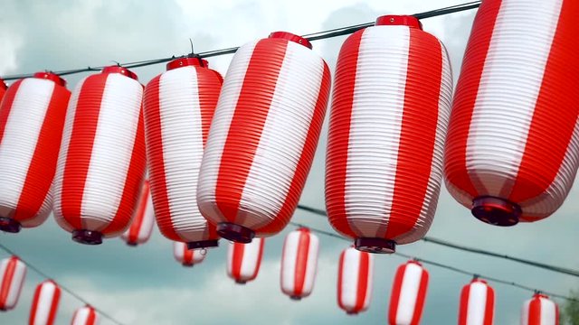 Many oriental paper red-white lanterns Chochin hanging on white cloudy blue sky background. Japanese lanterns hanging on bright sky background