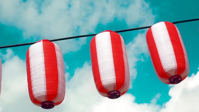 Oriental paper red-white lanterns Chochin hanging on white cloudy blue sky background. Japanese lanterns hanging on bright sky background. Pan left