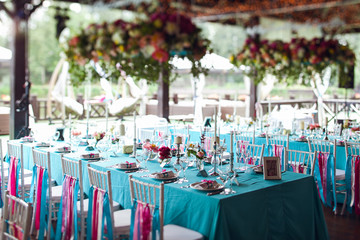 Table setting for a wedding party in pink and turquoise colors. White chairs with silk ribbons. Luxury decor. Plates, glasses, cutlery and candles. Vases with flowers.