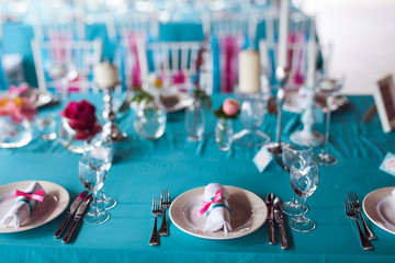 Table setting for a wedding party in pink and turquoise colors. White chairs with silk ribbons. Luxury decor. Plates, glasses, cutlery and candles. Vases with flowers.