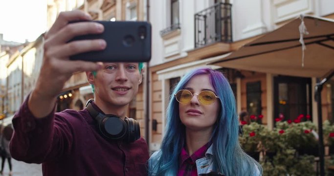 Caucasian young hipsters guy and girl with blue hair posing to the smartphone camera while taking selfie photos at the street in the town. Close up. Outside.
