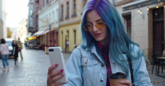 Close up of the Caucasian young stylish hipster girl with blue hair and in sunglasses taking selfie photos with cup of coffee to-go in hands on the smartphone at the street in the town. Outdoor.