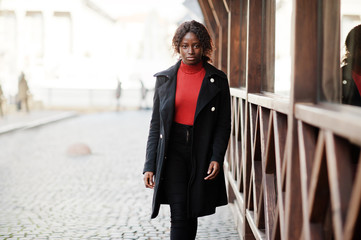 Obraz premium Portrait of a curly haired african woman wearing fashionable black coat and red turtleneck posing outdoor.