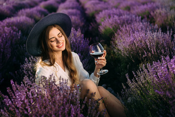 The girl in the hat sit in the middle of a lavender field with a glass of wine and closes her eyes