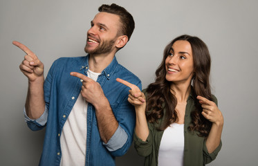 Look here! Close up photo of a magnificent couple in white t-shirts and colorful shirts, who are pointing to the left with their index fingers, looking in the same direction and smiling.