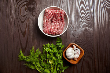 Fototapeta na wymiar Bowl with raw ground beef and condiments on wooden table, top view. Cooking minced meat with fresh seasonings, flat lay. White garlic heads and cloves, green dill and parsley leaves