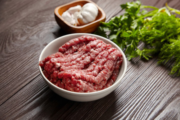 Bowl with raw ground beef and condiments on wooden table, closeup. Cooking minced meat with fresh seasonings. White garlic heads and cloves, green dill and parsley leaves