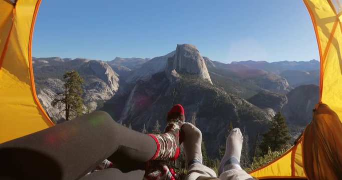Camping rest in Yosemite Valley. A couple lay inside the yellow tent and point their fingers at the famous Half Dome rock that is seen outside on a picturesque mountain ridge. Yosemite National Park
