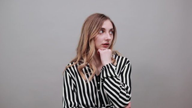 Boring woman in black and white striped shirt looking at camera, keeping fist over chin, keeping hand on stomach isolated on gray background in studio. People sincere emotions, lifestyle concept.