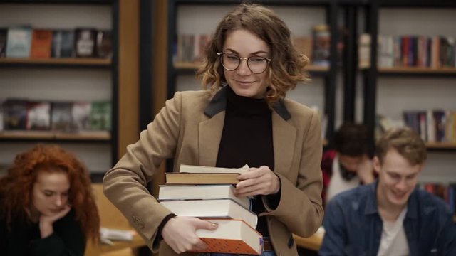 Slow motion footage of a girl in brown jacket and glasses walking by row with pile of books she took from the shelf. Other students sitting, studying in college, university library. Front view