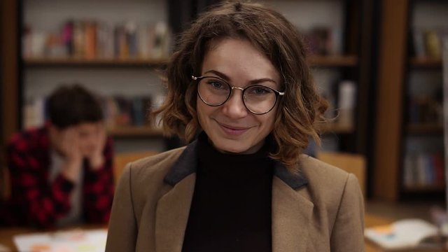 Portrait of an attractive short haired european girl student in glasses standing in high school library smiling looking at camera and smiling. Education, literature and people concept. Classmates on