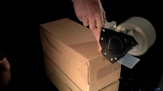 This video shows a brown box on a black background being closed, packaged, and taped with a shipping tape gun.