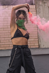 Beautiful young feminist activist dressed in black with face covered with a green bandana and holding light up colored red smoke bombs