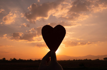 Fototapeta premium Silhouette of the heart shaped cardboard in hand and the sun shining through the heart on sunset background. Concepts of love and valentines day.