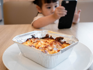 Little Asian child girl eating lunch while watching cartoon on the smartphone in a restaurant with her family. Child addicted to mobile phones.