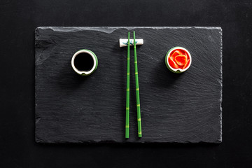 Table served for eating sushi. Chopsticks, small bowls with ginger and sause, mat on black background top-down