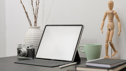 Close up view of blank screen tablet  with coffee cup, stylus, camera, wooden figure and decoration on black desk with white wall