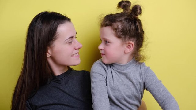 Cute kid daughter and mother bloggers talking to camera recording vlog together, young mom embracing little child girl waving hand shooting video blog making video call on background yellow wall