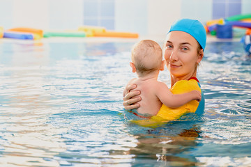 Young mother aged 30 years and baby son of 5 months are in pool. Smiling woman with beautiful snow-white smile is dressed in fashionable swimming suit swimsuit and cap. Free space for advertising