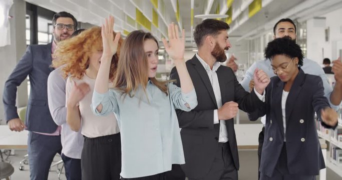 Excited office workers dancing, smiling , having good time in modern workspace. Front view of happy business team enjoying startup success. Concept of team win, victory, internationality.
