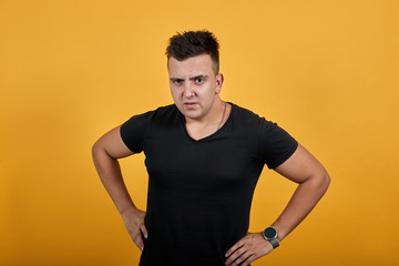 Caucasian young man keeping hands on belt, angry wearing black shirt isolated on orange background in studio. People sincere emotions, lifestyle concept.