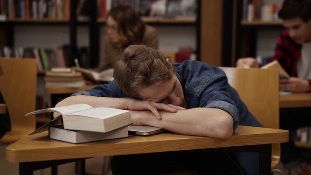 Young european student in a college library or class sleeping on desk with pile of books on it. Classmates and bookshelf on the background