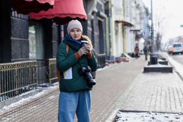 Girl in winter clothes with camera and map in the historical center of the city is holding a cup of coffee on the street