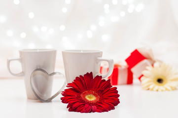 red gift boxes with ribbons, a pair of white cups and flowers on a white delicate background with bokeh.
