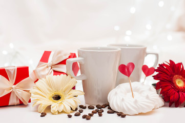 red gift boxes with ribbons, a pair of white cups and flowers on a white delicate background with bokeh.