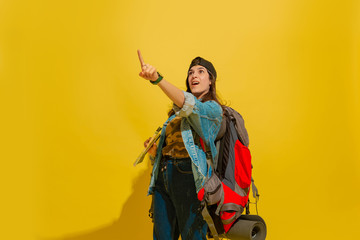 Fototapeta premium Astonished, looking up. Portrait of a cheerful young caucasian tourist girl with bag in jeans clothes isolated on yellow studio background. Preparing for traveling. Resort, human emotions, vacation.