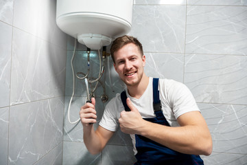 smiling man worker in uniform with screwdriver in one hand and showing like with another hand repairing boiler at home in toilette professional repair service