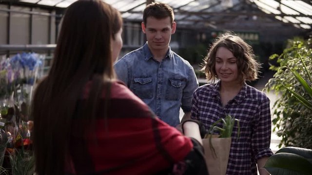 European young couple are recieving organic food in brown paper bag from female seller in greenhouse. A man paying with a american dollar bank note Cheerful, smillling woman selling organic food from
