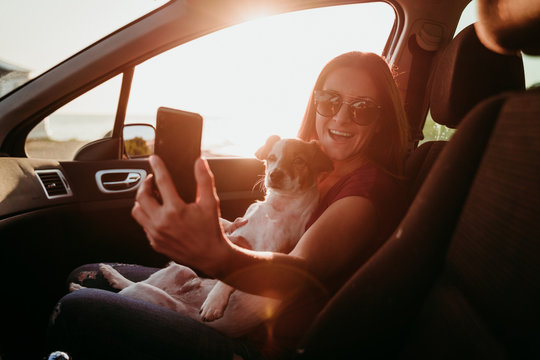 young woman and her cute dog in a car at sunset. travel concept. woman taking a selfie with mobile phone