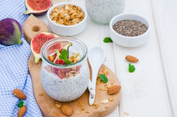 Chia pudding with yogurt, homemade granola, fresh figs and honey in a glass jar on a board on a white wooden background. Copy space.
