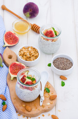 Chia pudding with yogurt, homemade granola, fresh figs and honey in a glass jar on a board on a white wooden background.