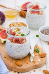 Chia pudding with yogurt, homemade granola, fresh figs and honey in a glass jar on a board on a white wooden background.