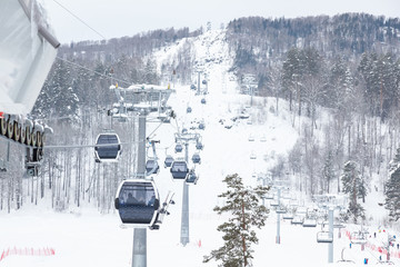 A gondola cableway with a booth suspended on a cable in which sits people with skis and snowboards on a background of mountains, trees with snow on winter. Ski resorts and snowboarding.
