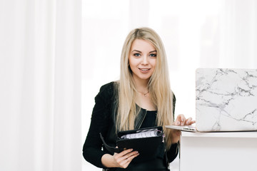 Beautiful smiling blonde girl with a notebook and pen in her hands, next to a laptop on a light background, office employee