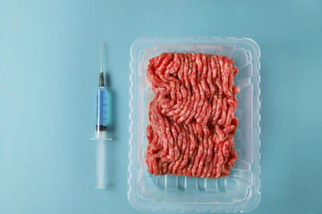 Ground beef in container and syringe with blue liquid – protein, antibiotic, hormones or other chemical and biological additives. Artificial cultured meat concept. Food of the future, top view, flat l