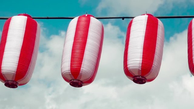 Oriental paper red-white lanterns Chochin hanging on white cloudy blue sky background. Japanese lanterns hanging on bright sky background. Pan left