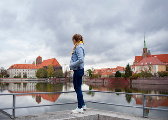  girl in a denim jacket stands with his back on the promenade and looks at the river