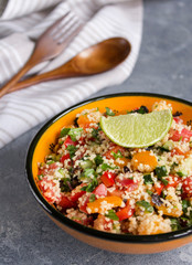 Tabouleh: vegetable couscous salad. Vegetables and couscous salad tabouleh in the orange ceramic bowl on concrete surface. Fresh summer salad with lime slice, wooden spoon, fork and white linen towel