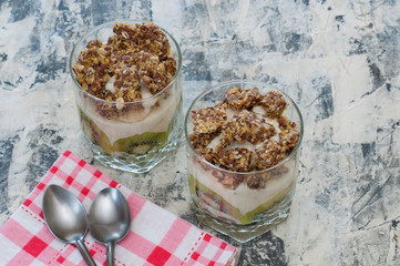 Dessert granola, Greek yogurt, kiwi and banana in two glass cups with spoons and pink checkered towel on gray concrete. Fitness, figure, body and healthy food