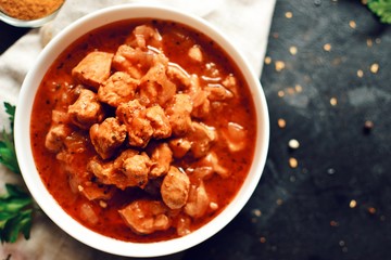 Tikka Masala. Chicken in tomato sauce in a white bowl. Spices and cherry tomatoes with parsley. Tasty chicken meat on a dark background. Horizontal view