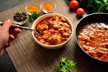 Indian food. Chicken Tikka Masala in a white bowl on a dark background. Appetizing meat with tomato sauce, gravy. Hands in the frame. Woman holds a spoon with food in her hand.