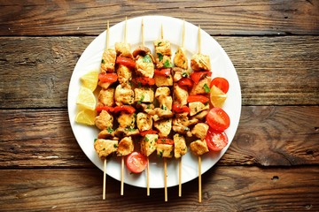Traditional greek kebab of souvlaki with tomatoes, parsley, onions, mushrooms and paprika. top view on a white plate. Chicken meat on wooden skewers. Wooden background texture. Horizontal view 