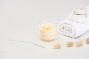 Fototapeta na wymiar fluffy bunny tail grass near burning white candle in glass and rolled towel with stones on marble white surface