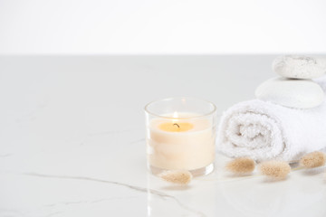 fluffy bunny tail grass near burning white candle in glass and rolled towel with stones on marble white surface