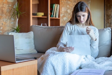 Serious young brunette woman looking at a paper sitting on the sofa at home with a blanket