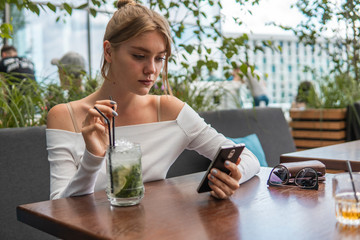 Cheerful blond woman blogger with cocktail is reading incoming sms message on smartphone connected to free wifi in cafe. Positive young student watching broadcasting online on modern mobile phone.